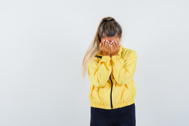Foto gratuita mujer rubia que cubre el rostro con las manos en chaqueta de bombardero amarillo y pantalón negro y mirando acosado