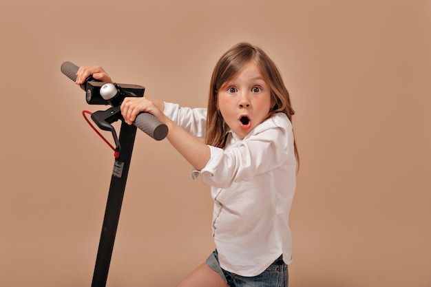 Foto gratuita niña adorable divertida con camisa blanca con scooter eléctrico posando con emociones sorpresa
