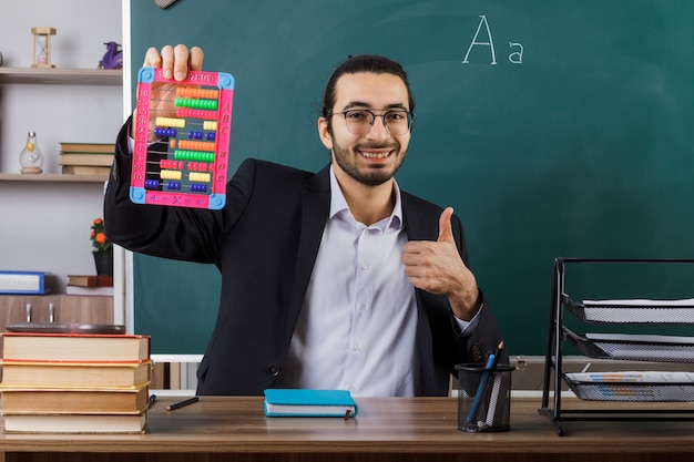 Foto gratuita sonriendo mostrando el pulgar hacia arriba profesor con gafas sosteniendo abacus sentado a la mesa con herramientas escolares en el aula