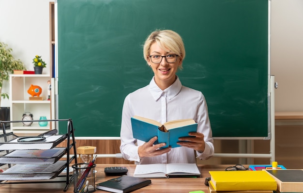 Foto gratuita sonriente joven rubia maestra con gafas sentados frente al escritorio con útiles escolares en el aula sosteniendo el libro abierto mirando al frente