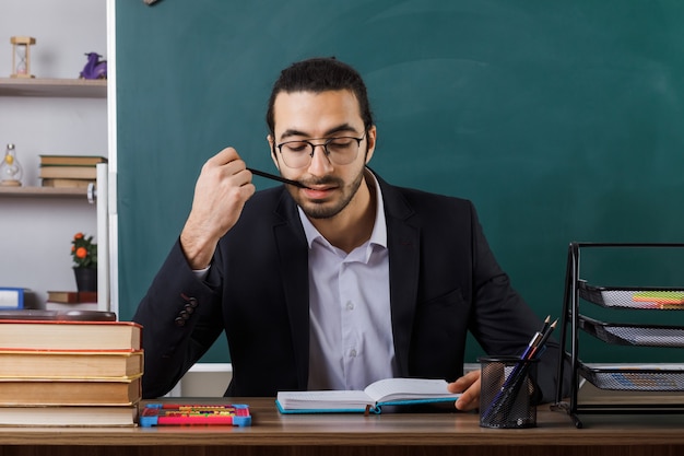 Foto gratuita profesor de sexo masculino complacido con gafas leyendo libro poniendo lápiz en la boca sentado a la mesa con herramientas escolares en el aula