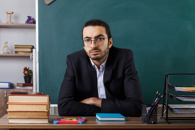 Foto gratuita profesor de sexo masculino confiado con gafas sentado a la mesa con herramientas escolares en el aula