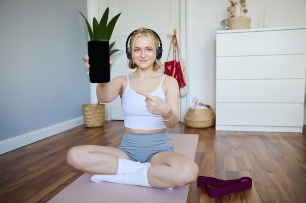 Foto gratuita retrato de una instructora de fitness sonriente, en forma y sana en casa señalando con el dedo y mostrándola