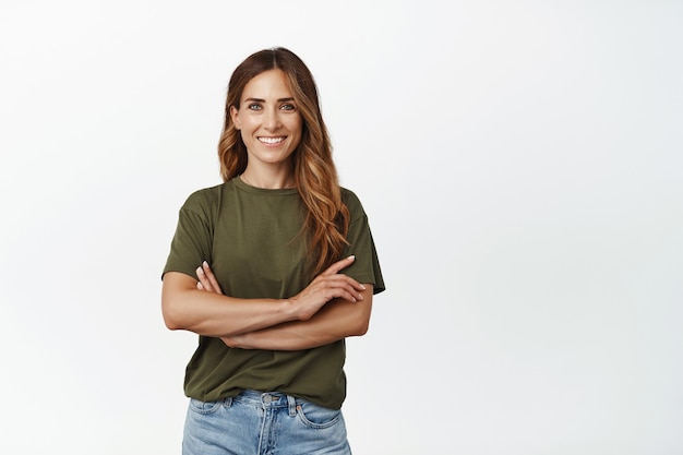 Foto gratuita retrato de mujer adulta confiada en camiseta verde, de pie con los brazos cruzados sobre el pecho y una sonrisa motivada y segura de sí misma, mirando al frente, de pie contra la pared blanca.
