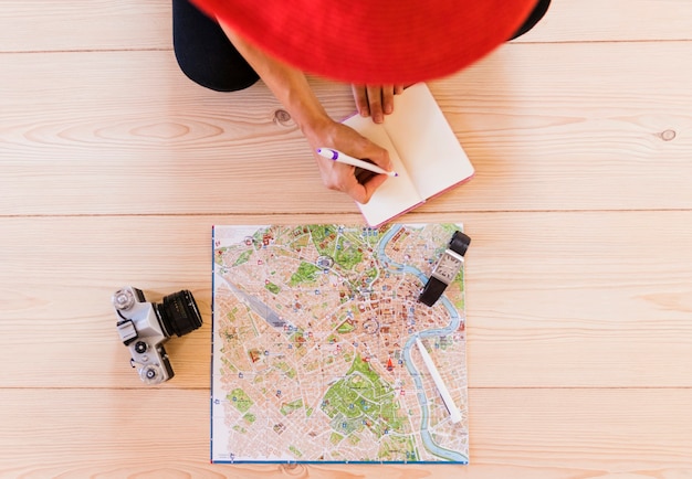 Foto gratuita vista elevada de la persona que escribe en el diario con el mapa; reloj de pulsera y cámara en mesa de madera.