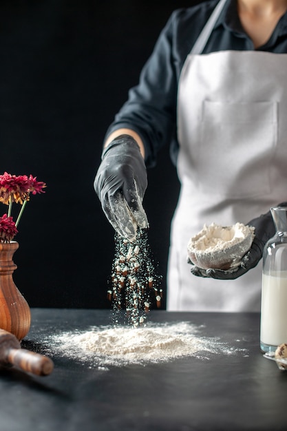 Vista frontal cocinera vertiendo harina blanca sobre la mesa para la masa en un trabajo oscuro pastel de pastelería panadería cocinar masa hornear pastel
