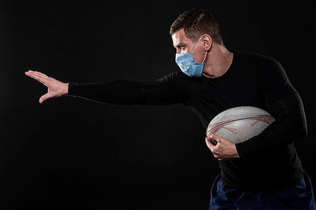 Vista lateral del jugador de rugby masculino con máscara médica y pelota