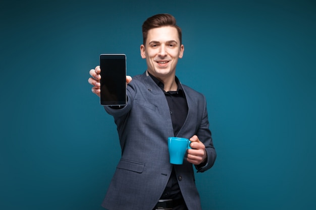 Il giovane uomo d'affari attraente in rivestimento grigio e camicia nera tiene la tazza blu e mostra il telefono