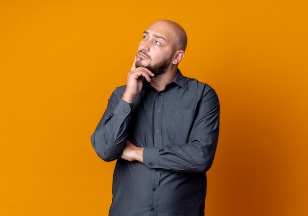 Free photo thoughtful young bald call center man putting hand on chin and looking at side isolated on orange wall