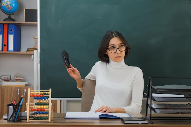 Бесплатное фото Думает, глядя в сторону, молодая учительница в очках, держащая калькулятор, сидящая за партой со школьными инструментами в классе