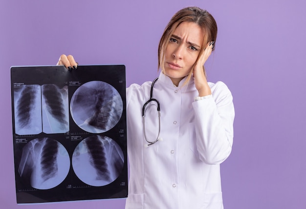 Бесплатное фото Усталая молодая женщина-врач в медицинском халате со стетоскопом держит рентгеновский снимок, положив руку на голову, изолированную на фиолетовой стене