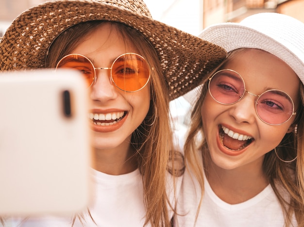 Бесплатное фото 2 молодых усмехаясь женщины битника белокурых в одеждах футболки лета белых. девушки, принимающие selfie автопортрет фотографий на смартфоне. ,