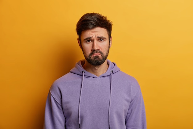 Free photo unamused sad miserable guy purses lips, frowns face with dissatisfaction, wears hoodie, poses indoor against yellow wall, sulks troubled as someone hurt his feelings, wears purple hoodie