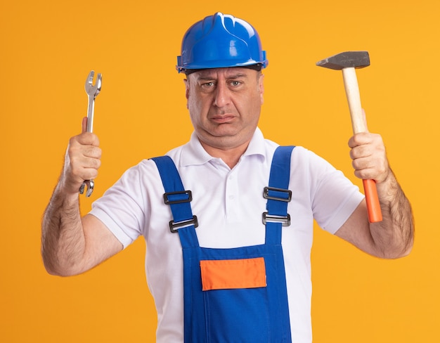 Бесплатное фото Недовольный взрослый строитель в униформе держит гаечный ключ и молоток, изолированные на оранжевой стене