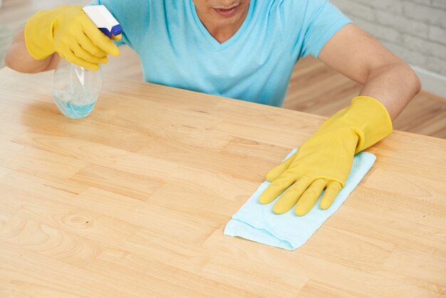 Неузнаваемый мужчина в резиновых перчатках опрыскивает стол и чистит тряпкой