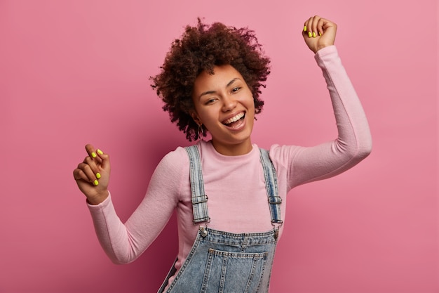 Upbeat dark skinned African American female model dances and feels joyful, wears turtleneck and denim sarafan, smiles broadly, has fun and moves with rhythm of music, isolated over pink wall