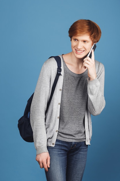 Бесплатное фото Вертикальный портрет молодой красивый студент с рыжими волосами, носить случайный наряд и рюкзак, улыбаясь, разговаривая по смартфон с другом по дороге в университет.