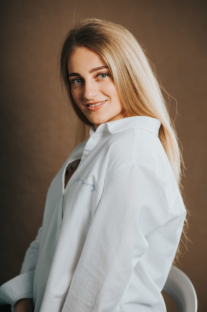 Бесплатное фото Вертикальный снимок привлекательной блондинки в белой рубашке, позирующей на коричневой стене