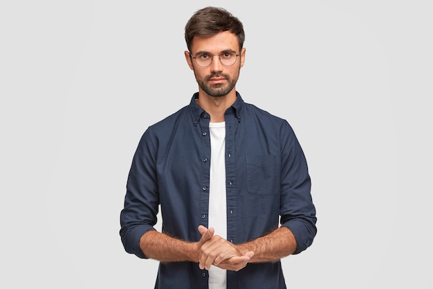 Free photo waist up portrait of handsome serious unshaven male keeps hands together, dressed in dark blue shirt, has talk with interlocutor, stands against white wall. self confident man freelancer
