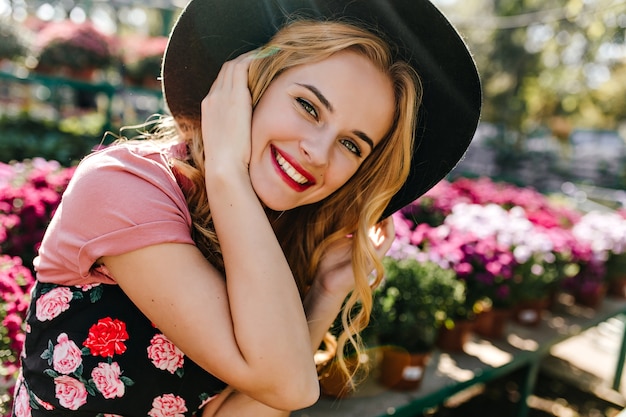Free photo white enchanting woman expressing happiness during portraitshoot with flowers. portrait of charming caucasian model in hat sitting in nature