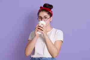 Бесплатное фото Обаятельная молодая счастливая красивая женщина в красной повязке для волос и белой повседневной футболке позирует на фоне лилового пространства, пьет кофе в бумажном стаканчике