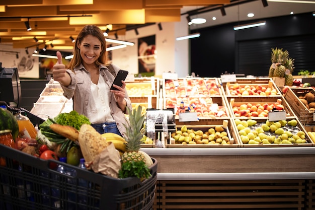 Free photo woman with smart phone in supermarket standing by the shelves full of fruit at grocery store holding thumbs up