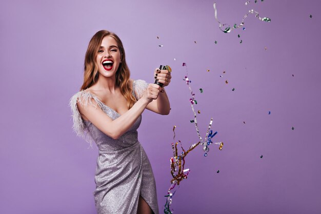 Женщина в праздничном платье бросает конфетти на изолированный фон Веселая девушка в серебряном наряде смеется на фиолетовом фоне во время вечеринки