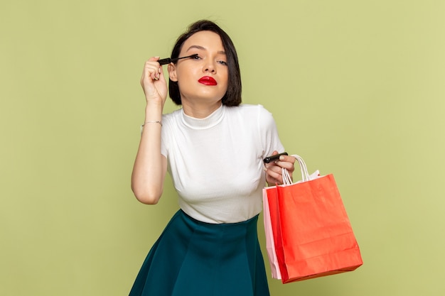 Бесплатное фото Женщина в белой блузке и зеленой юбке держит пакеты с покупками и делает макияж