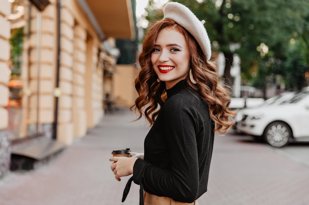 Бесплатное фото Замечательная рыжая женщина в берете, глядя через плечо. длинноволосая улыбающаяся девушка пьет кофе на улице.