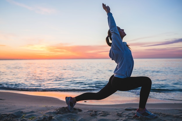 Бесплатное фото Молодая женщина делает спортивные упражнения на пляже восхода солнца утром