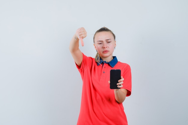 Free photo young woman looking at mobile phone with thumb down in t-shirt and looking upset