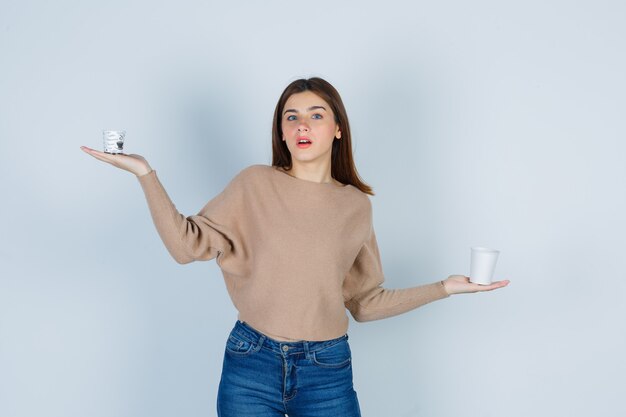 Бесплатное фото Молодая женщина разводит ладони с бумажными стаканчиками в свитере, джинсах и удивленно смотрит, вид спереди.