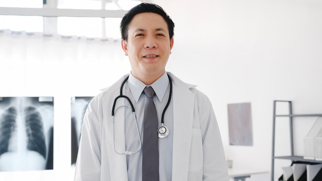 Foto gratuita giovane medico maschio asiatico in uniforme medica bianca con stetoscopio che guarda l'obbiettivo, sorriso e braccia incrociate durante la videoconferenza con il paziente in ospedale.
