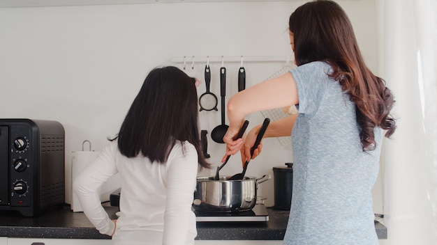 Молодая азиатская японская мама и дочь варя дома. Женщины образа жизни счастливые делая макаронные изделия и спагетти совместно для еды завтрака в современной кухне на доме в утре.