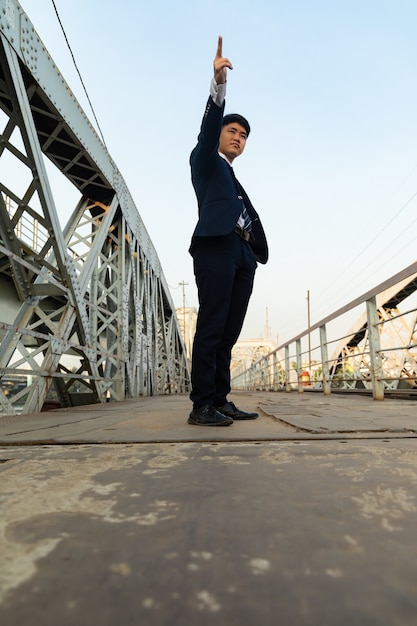 Free photo young asian man in a suit standing on a bridge while pointing up