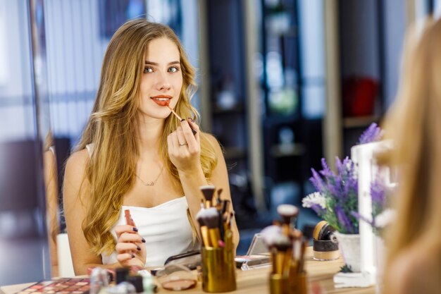 Молодая красивая девушка с длинными светлыми волосами рисует губы красной помадой перед зеркалом