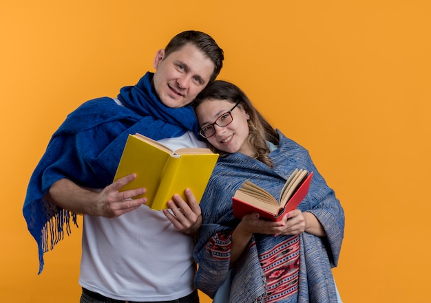 Free photo young couple man and woman with blankets holding books happy and positive smiling standing together over orange wall