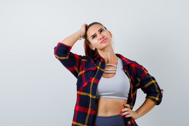Free photo young female in crop top, checkered shirt, pants with hand on head while keeping hand on hip and looking pensive , front view.