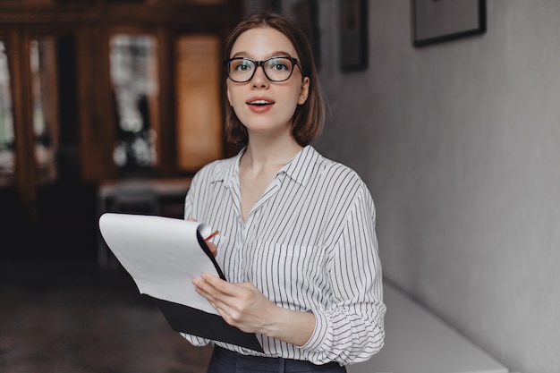 Бесплатное фото Молодая сотрудница в полосатой блузке держит папку с документами и смотрит в камеру через очки.