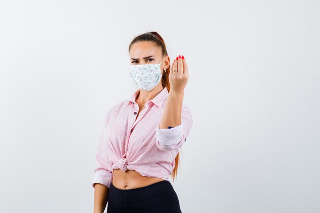 Young female showing Italian gesture in shirt, pants, medical mask and looking delighted. front view.