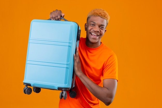 Free photo young handsome boy wearing orange t-shirt holding travel suitcase smiling cheerfully positive and happy standing over orange wall