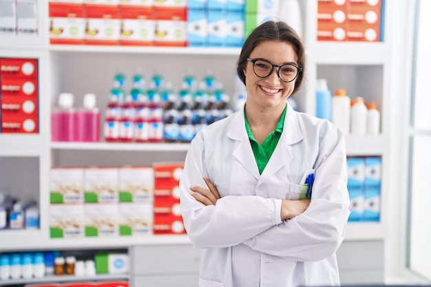 Бесплатное фото Молодая латиноамериканка-фармацевт уверенно улыбается, стоя со скрещенными руками в аптеке