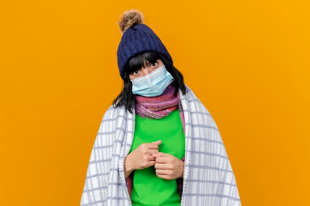 Бесплатное фото Молодая больная кавказская девушка в зимней шапке и шарфе с маской, завернутой в клетчатые кулаки, изолированной на оранжевой стене с копией пространства