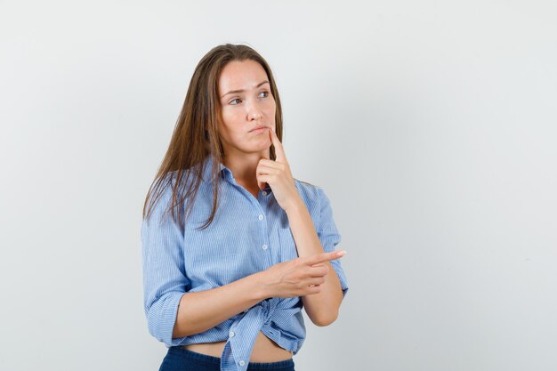 Young lady pointing to side in blue shirt, pants and looking pensive. front view.
