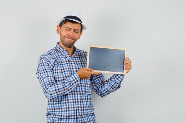 Young man in checked shirt, hat holding blank frame with closed eyes