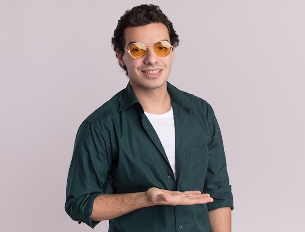 Бесплатное фото Молодой человек в зеленой рубашке в очках смотрит вперед с улыбкой на лице, представляя что-то рукой, стоящей над белой стеной