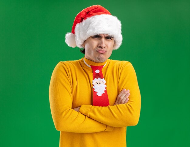 Young man in yellow turtleneck and santa hat with funny tie looking at camera displeased making wry mouth with disappointed expression standing over green background