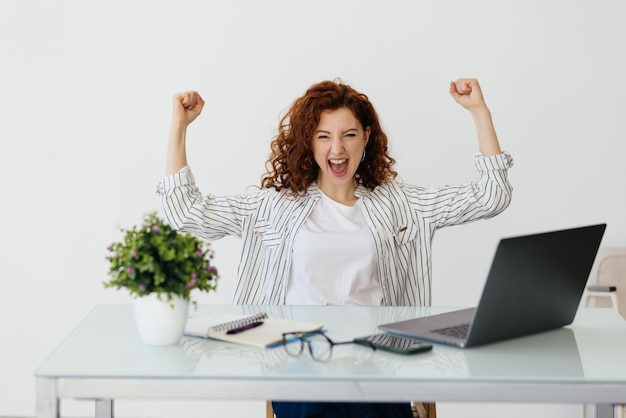 Бесплатное фото Молодая рыжеволосая кудрявая женщина, работающая со своим ноутбуком, поднимает кулак после концепции победителя