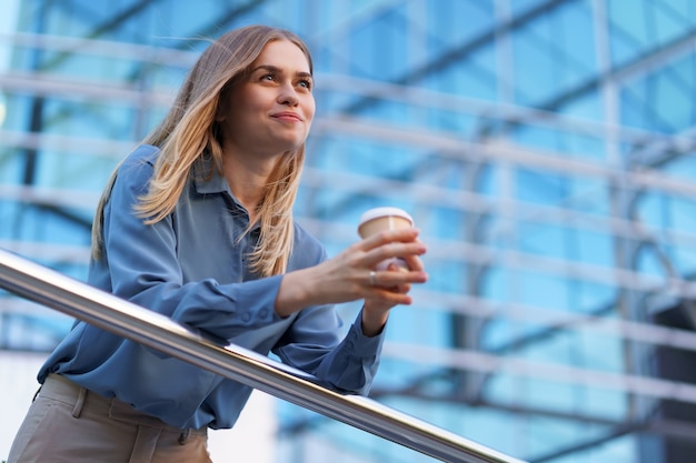 Бесплатное фото Молодая улыбающаяся профессиональная женщина, имеющая перерыв на кофе в течение ее полного рабочего дня. она держит бумажный стаканчик на открытом воздухе возле бизнес-здания, расслабляясь и наслаждаясь напитком.
