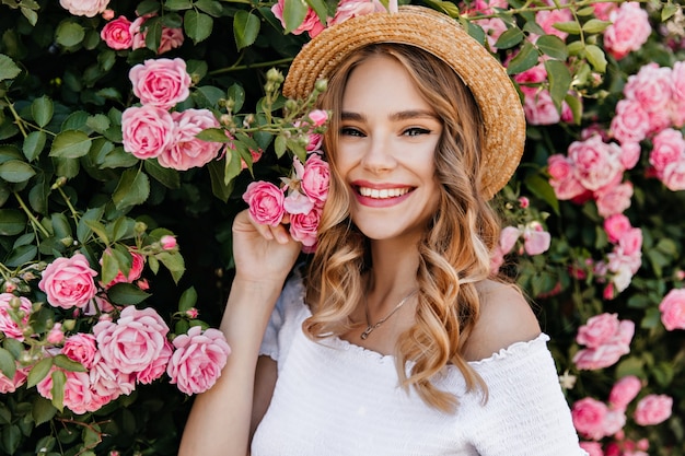Free photo adorable girl with curly blonde hair posing in garden. portrait of caucasian glad woman holding pink flower.
